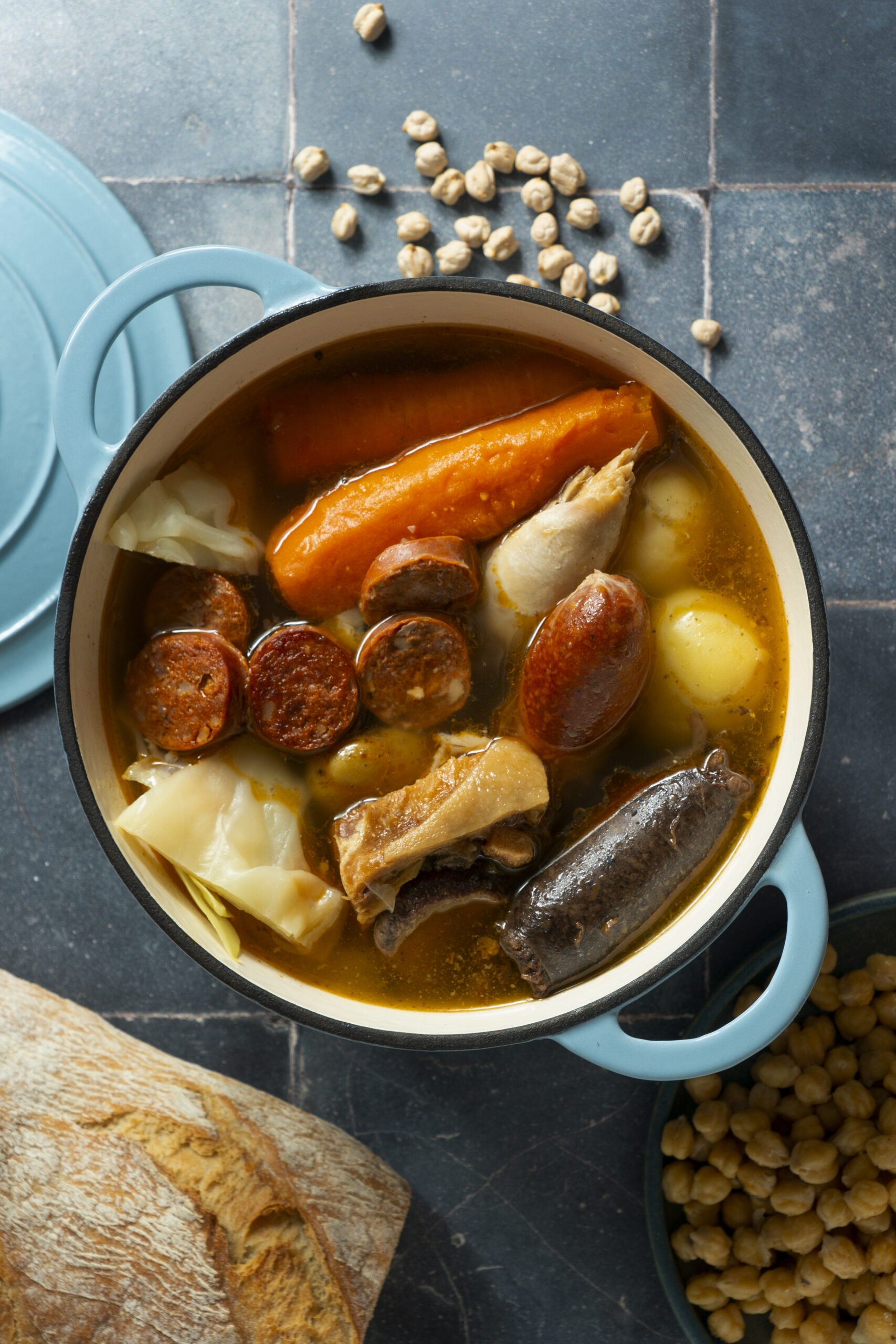 Plat mijoté traditionnel dans une cocotte en fonte bleue contenant carottes, pommes de terre, chou, pois chiches, morceaux de viande et différentes saucisses, accompagné de pain rustique. Vue de dessus d’un repas chaleureux et authentique.